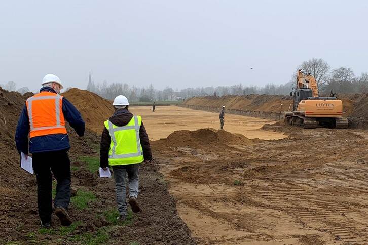 Afbeelding van een archeologische opgraving met op de voorgrond twee personen in veiligheidskleding en op de achtergrond een graafmachine en de opgraving